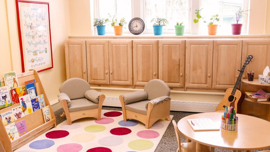 Cosy library nook in a classroom with a colourful spot placement mat, low book shelf, two comfy chairs, and natural light coming in from window.